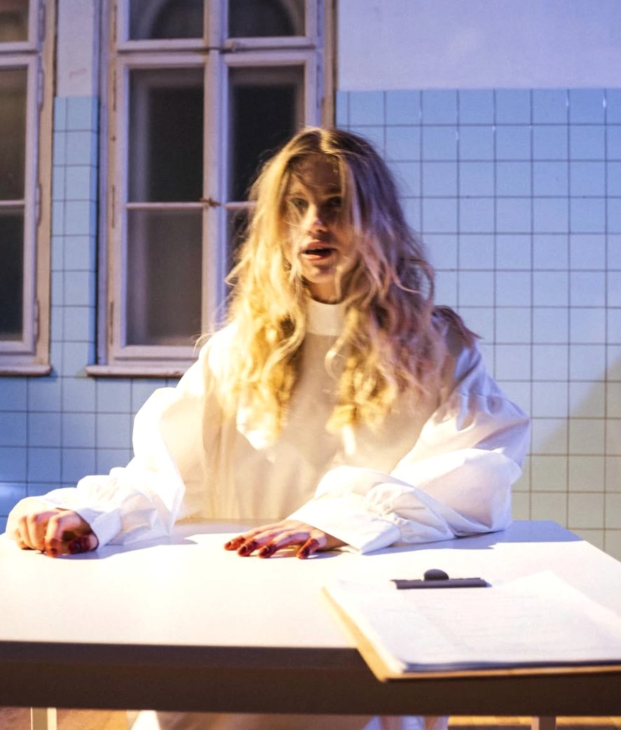A figure in white at a table, blood on the hands, a stark gaze